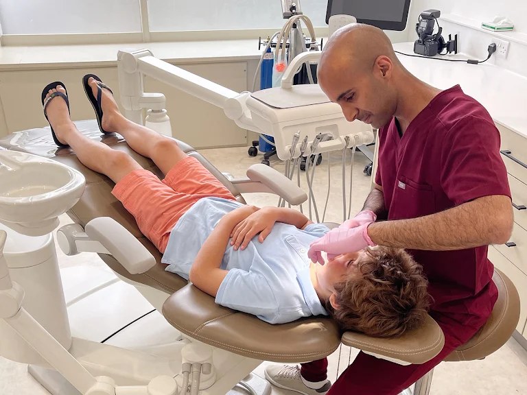Dr Kunal Patel in burgundy scrubs leaning over a young patient lying back in the dental chair, the child relaxed with arms folded.
